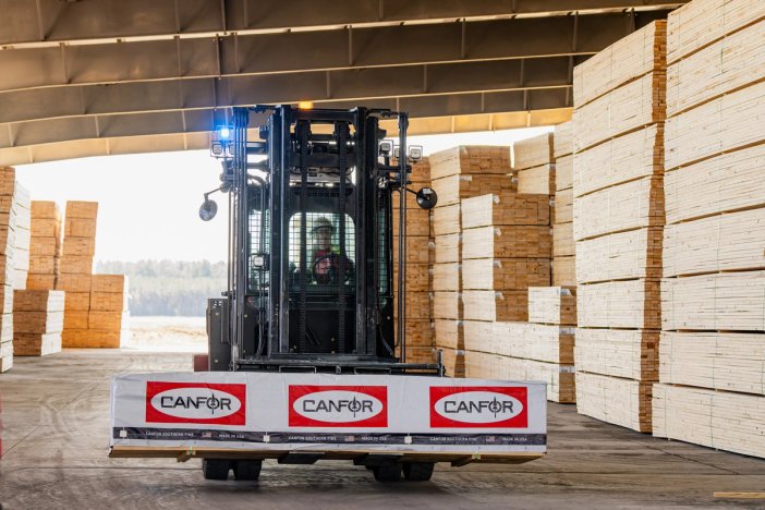 A forklift carries Canfor branded lumber through a large warehouse filled with tall stacks of wood.
