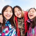 three young Indigenous women are smiling and laughing, posing for a picture in the snow .