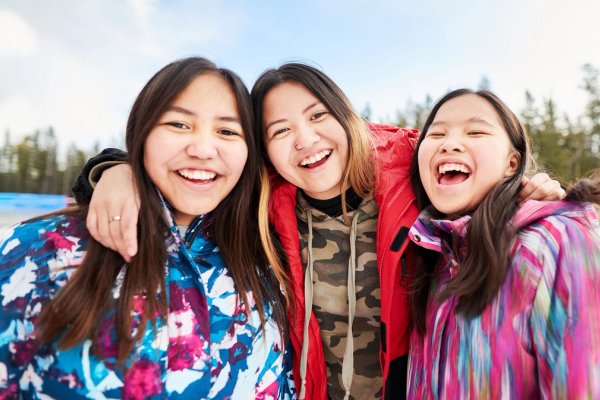 three young Indigenous women are smiling and laughing, posing for a picture in the snow .