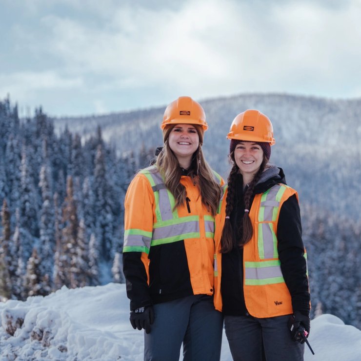 two women wearing hard hats and vests are standing next to each other in the snow .
