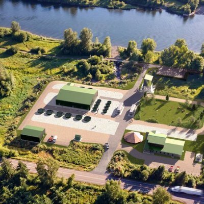 Aerial view of a modern facility with green buildings and circular tanks next to a river, surrounded by trees.
