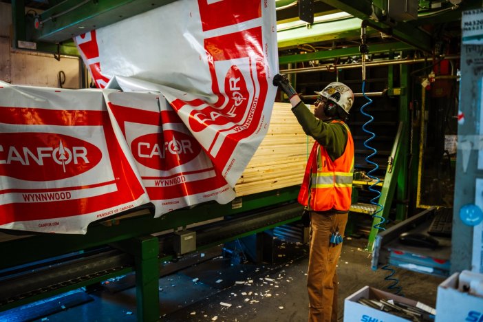 a man in an orange vest is standing next to a machine that is wrapping finished lumber .