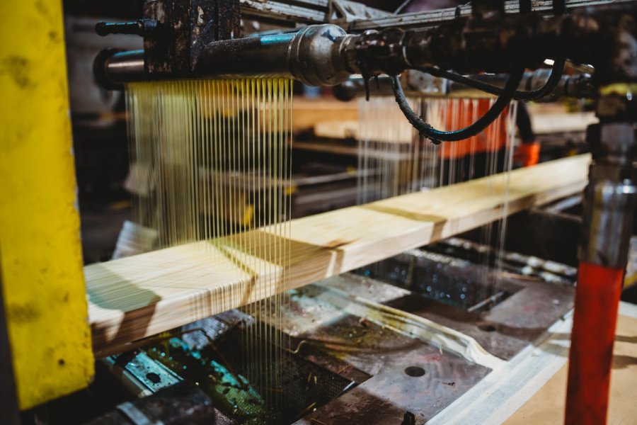 Industrial machinery applies thin, yellow streams of adhesive onto a wooden board.