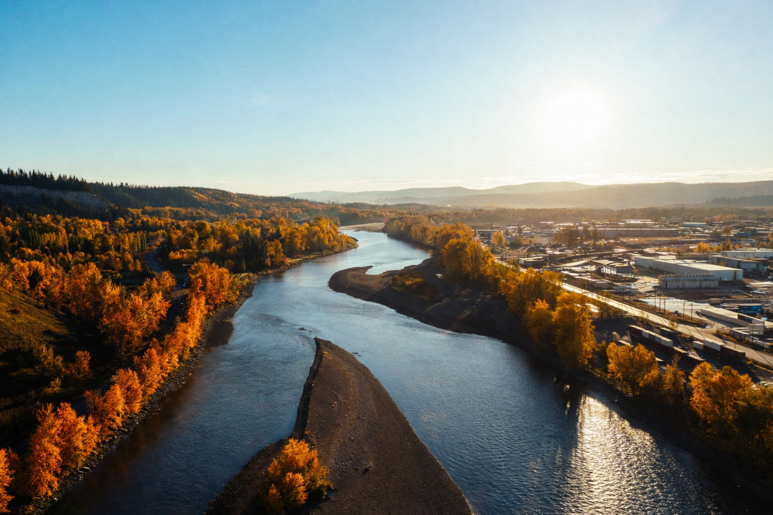 A winding river flows between vibrant orange autumn trees and a town under golden sunlight.