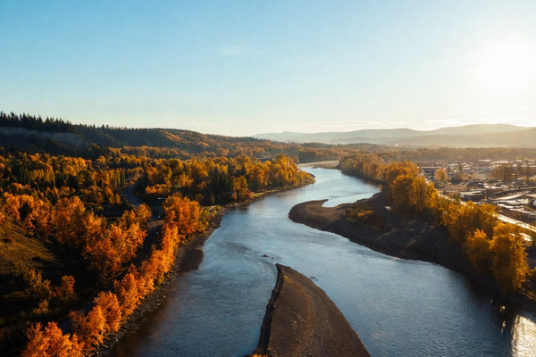 A winding river flows between vibrant orange autumn trees and a town under golden sunlight.