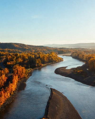 A winding river flows between vibrant orange autumn trees and a town under golden sunlight.