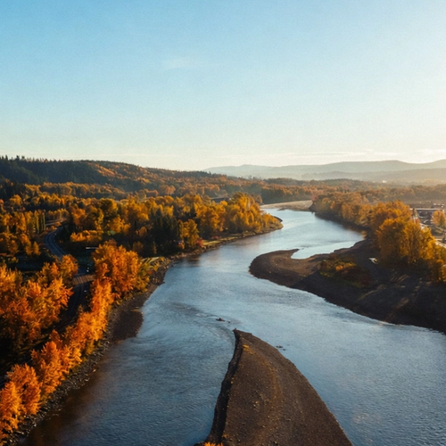 A winding river flows between vibrant orange autumn trees and a town under golden sunlight.