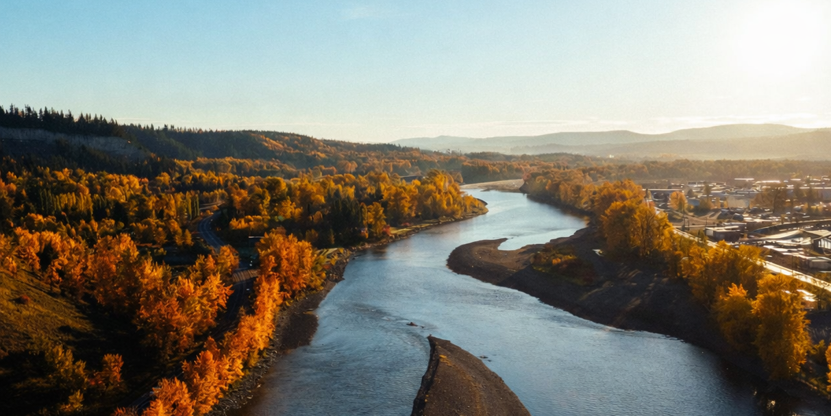 A winding river flows between vibrant orange autumn trees and a town under golden sunlight.