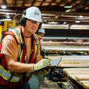 a man in safety vest, hard hat, glasses and gloves is standing next to a conveyor line moving lumber in a sawmill. he is holding a notebook and taking notes.