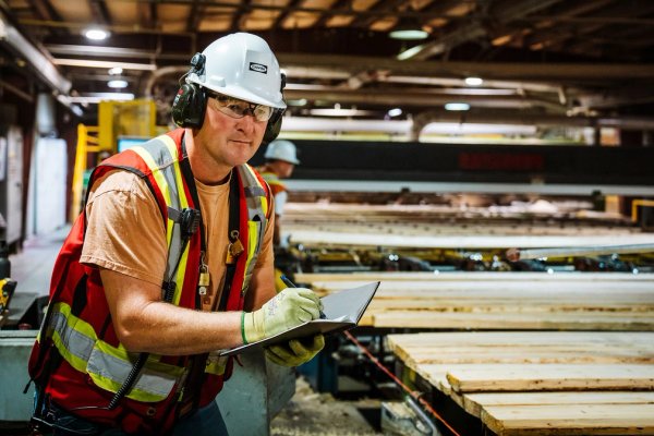 a man in safety vest, hard hat, glasses and gloves is standing next to a conveyor line moving lumber in a sawmill. he is holding a notebook and taking notes.