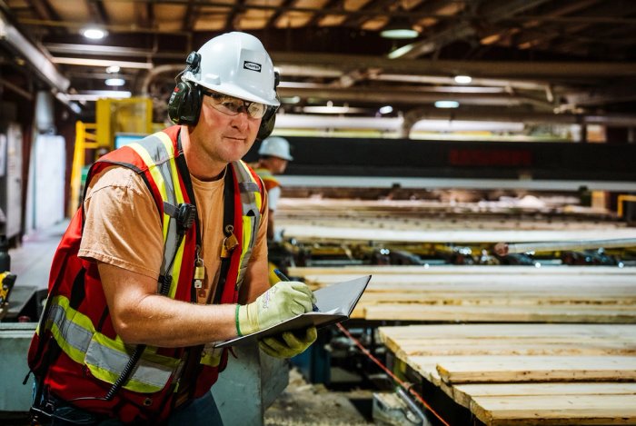 a man in safety vest, hard hat, glasses and gloves is standing next to a conveyor line moving lumber in a sawmill. he is holding a notebook and taking notes.