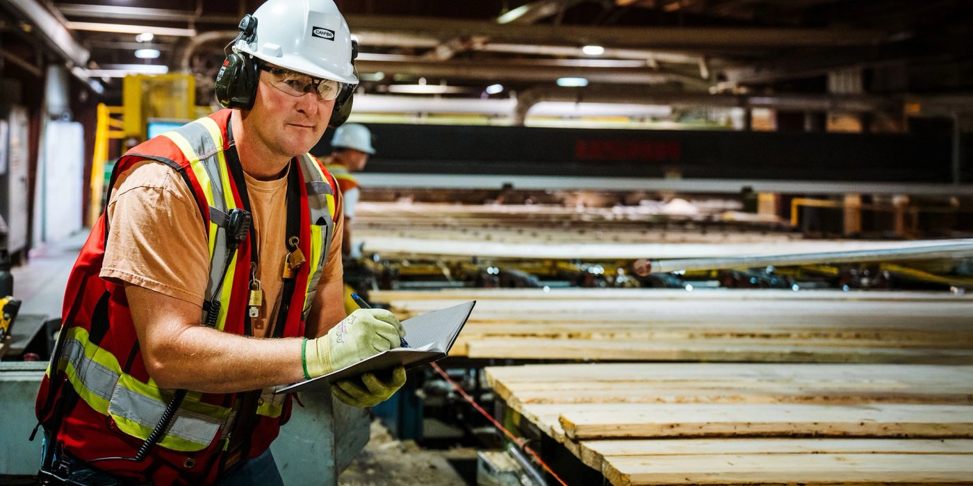 a man in safety vest, hard hat, glasses and gloves is standing next to a conveyor line moving lumber in a sawmill. he is holding a notebook and taking notes.