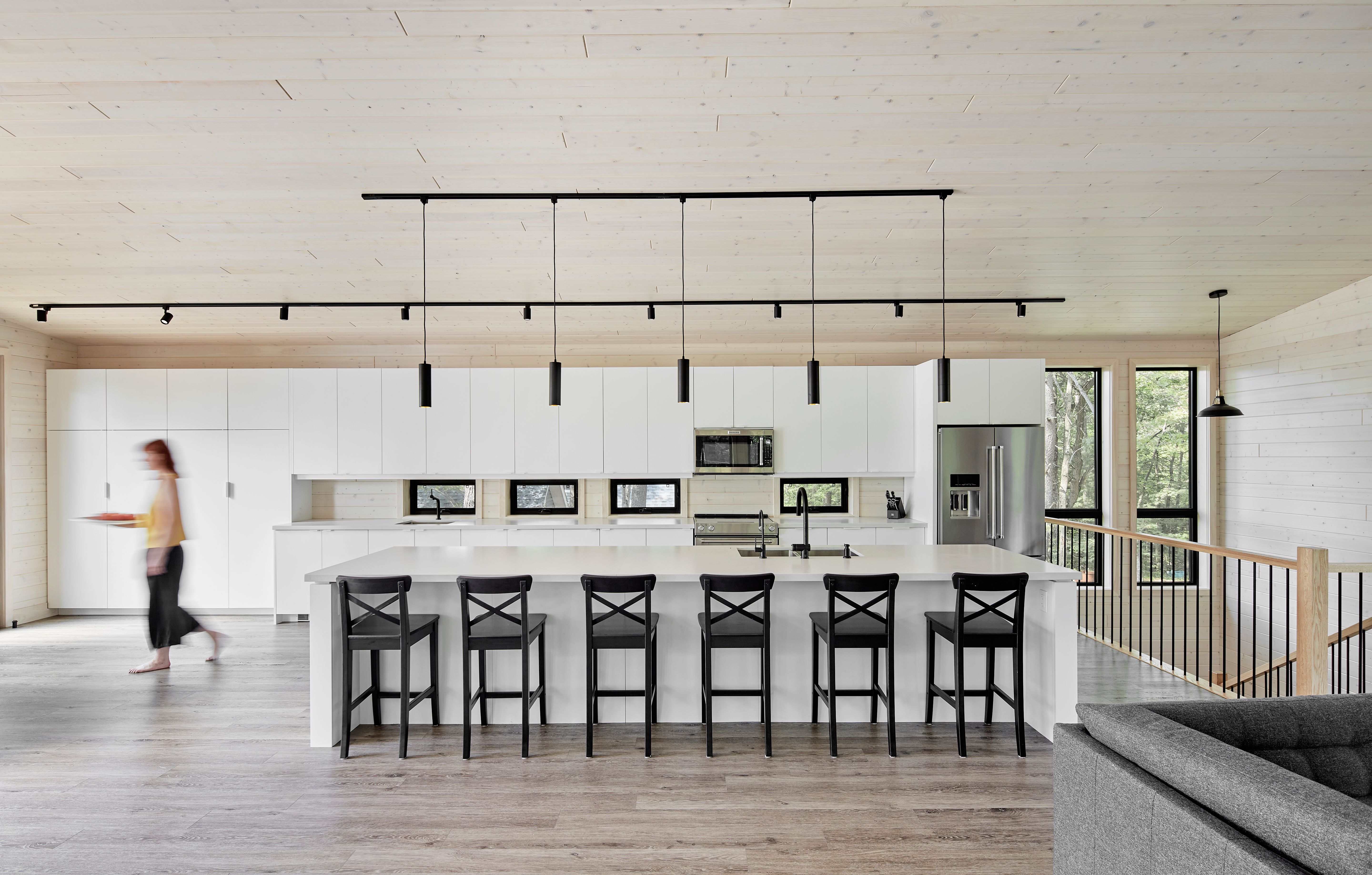 A modern kitchen with white cabinets, a large island with black bar stools, a blurred person walking, and a light wood ceiling.