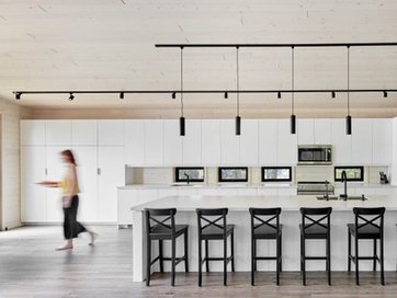 A modern kitchen with white cabinets, a large island with black bar stools, a blurred person walking, and a light wood ceiling.