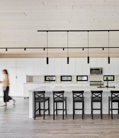 White modern kitchen with a large island, black stools, tall windows with a forest view, and a blurred person walking.