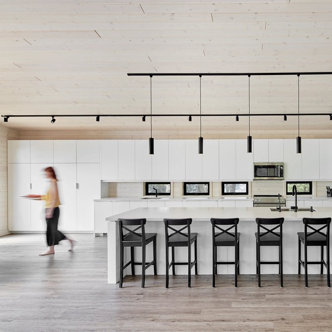 A modern kitchen with white cabinets, a large island with black bar stools, a blurred person walking, and a light wood ceiling.