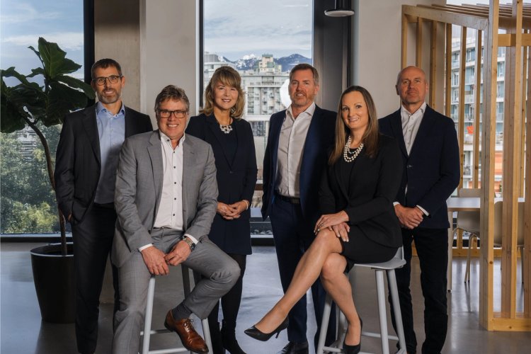 A group of six professionals, five men and one woman, in business attire posing in an office with a city view.