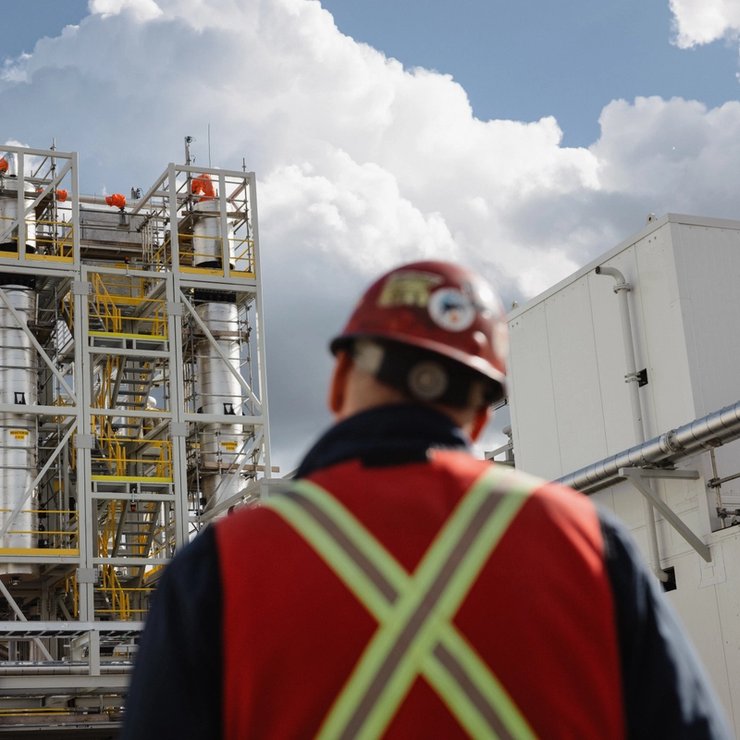 a man wearing a hard hat and safety vest is standing in front of a large industrial plant .