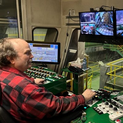 a man in a plaid shirt is sitting at a control panel in front of three monitors .
