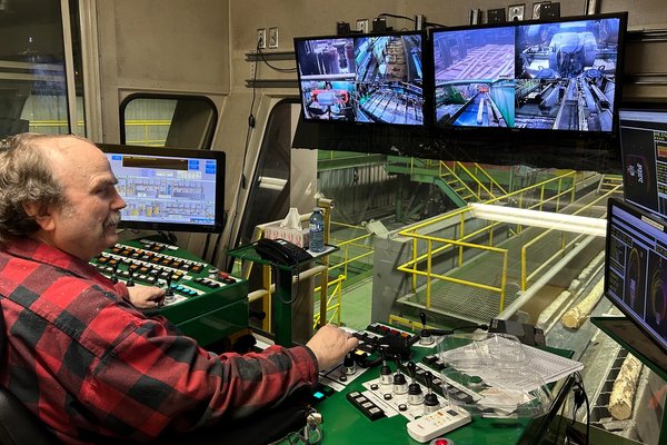 a man in a plaid shirt is sitting at a control panel in front of three monitors .