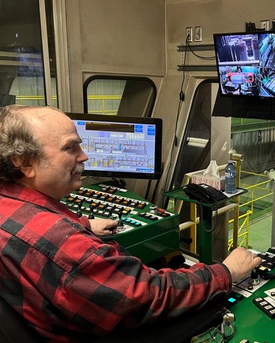 a man in a plaid shirt is sitting at a control panel in front of three monitors .