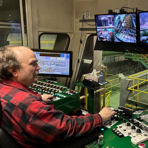 a man in a plaid shirt is sitting at a control panel in front of three monitors .