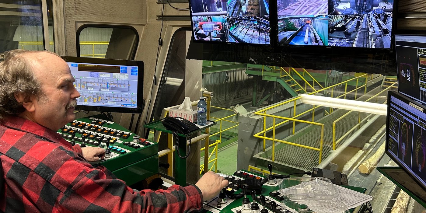 a man in a plaid shirt is sitting at a control panel in front of three monitors .