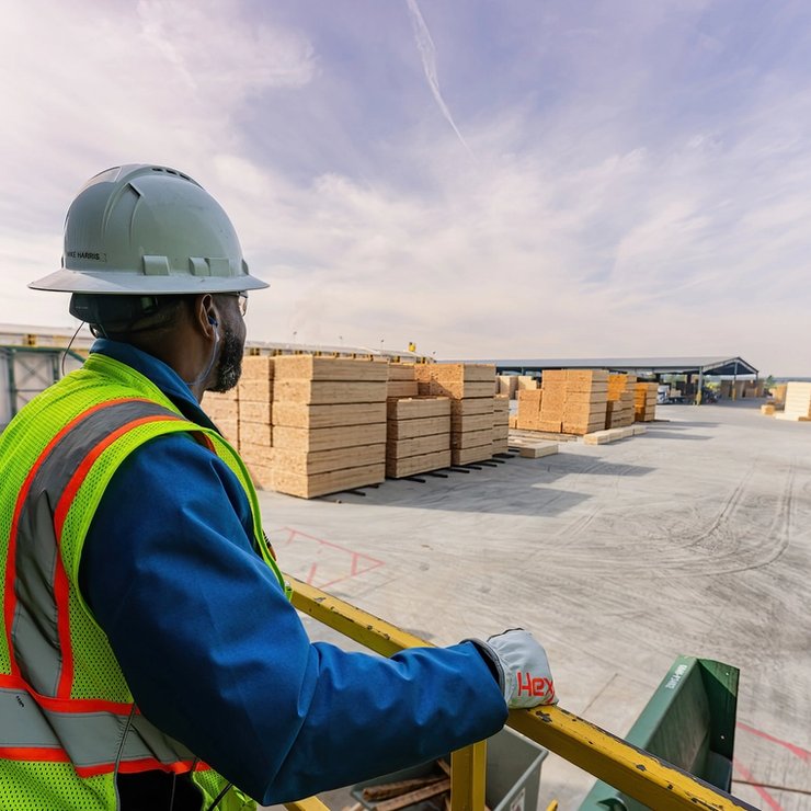 An employee wearing a hard hat and safety vest is standing overlooking a sawmill property.