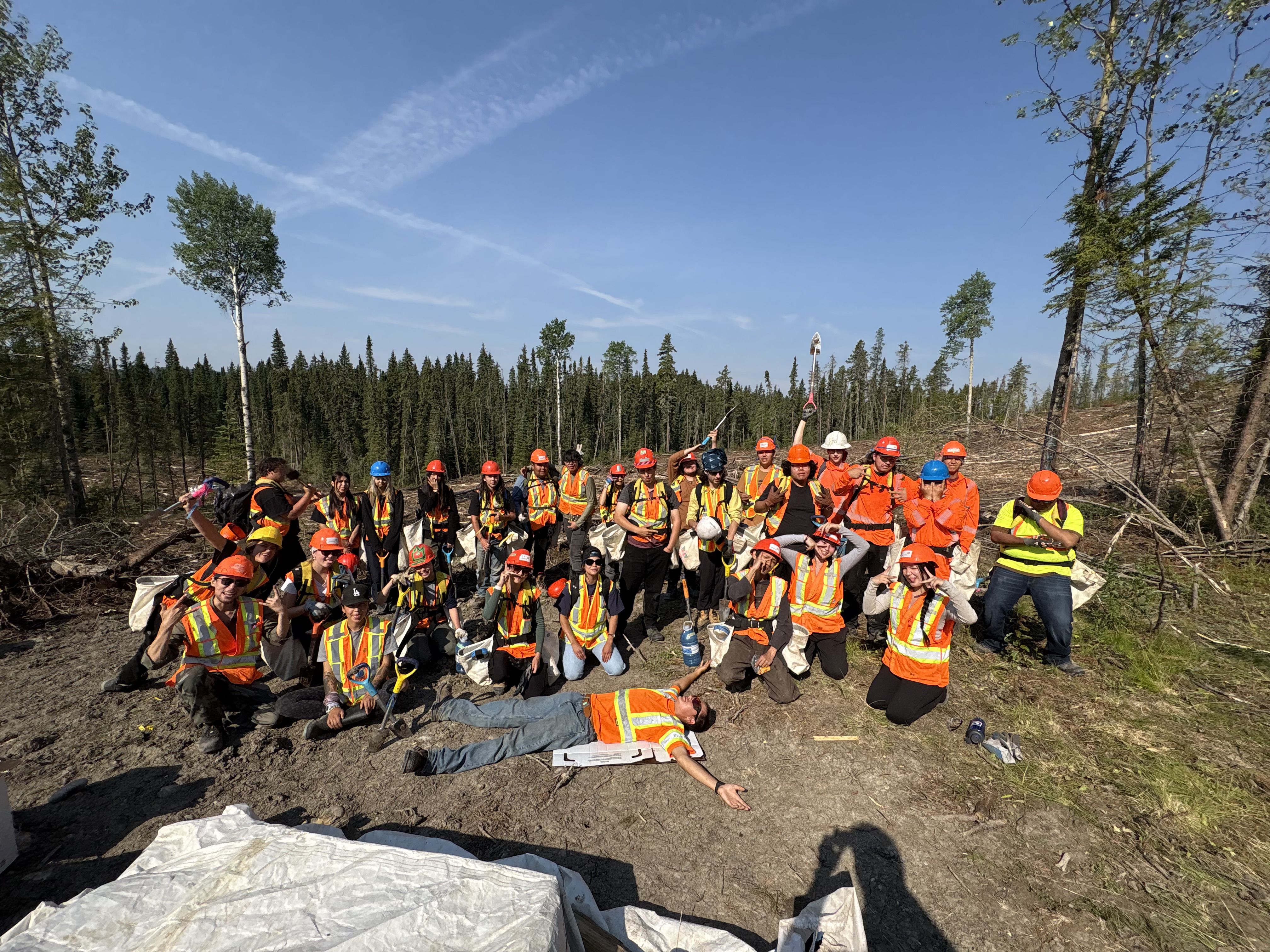 A team of two dozen workers in hard hats and high-visibility vests pose in a deforested area under a blue sky, with one worker lying on the ground.