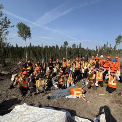 A team of two dozen workers in hard hats and high-visibility vests pose in a deforested area under a blue sky, with one worker lying on the ground.