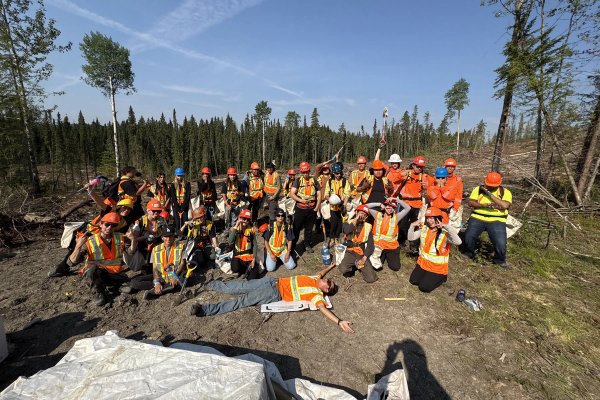 A team of two dozen workers in hard hats and high-visibility vests pose in a deforested area under a blue sky, with one worker lying on the ground.