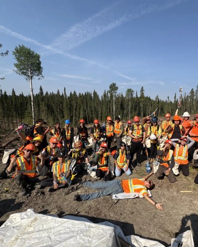 A team of two dozen workers in hard hats and high-visibility vests pose in a deforested area under a blue sky, with one worker lying on the ground.