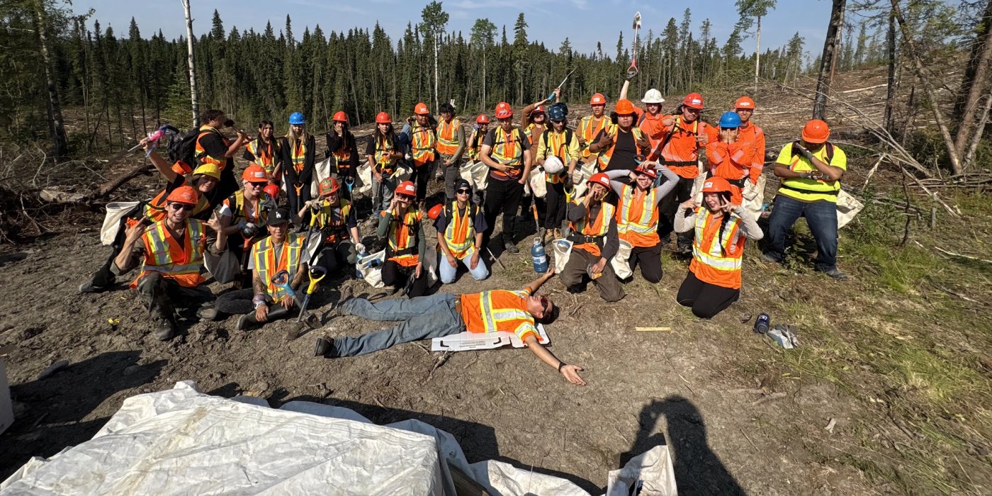A team of two dozen workers in hard hats and high-visibility vests pose in a deforested area under a blue sky, with one worker lying on the ground.