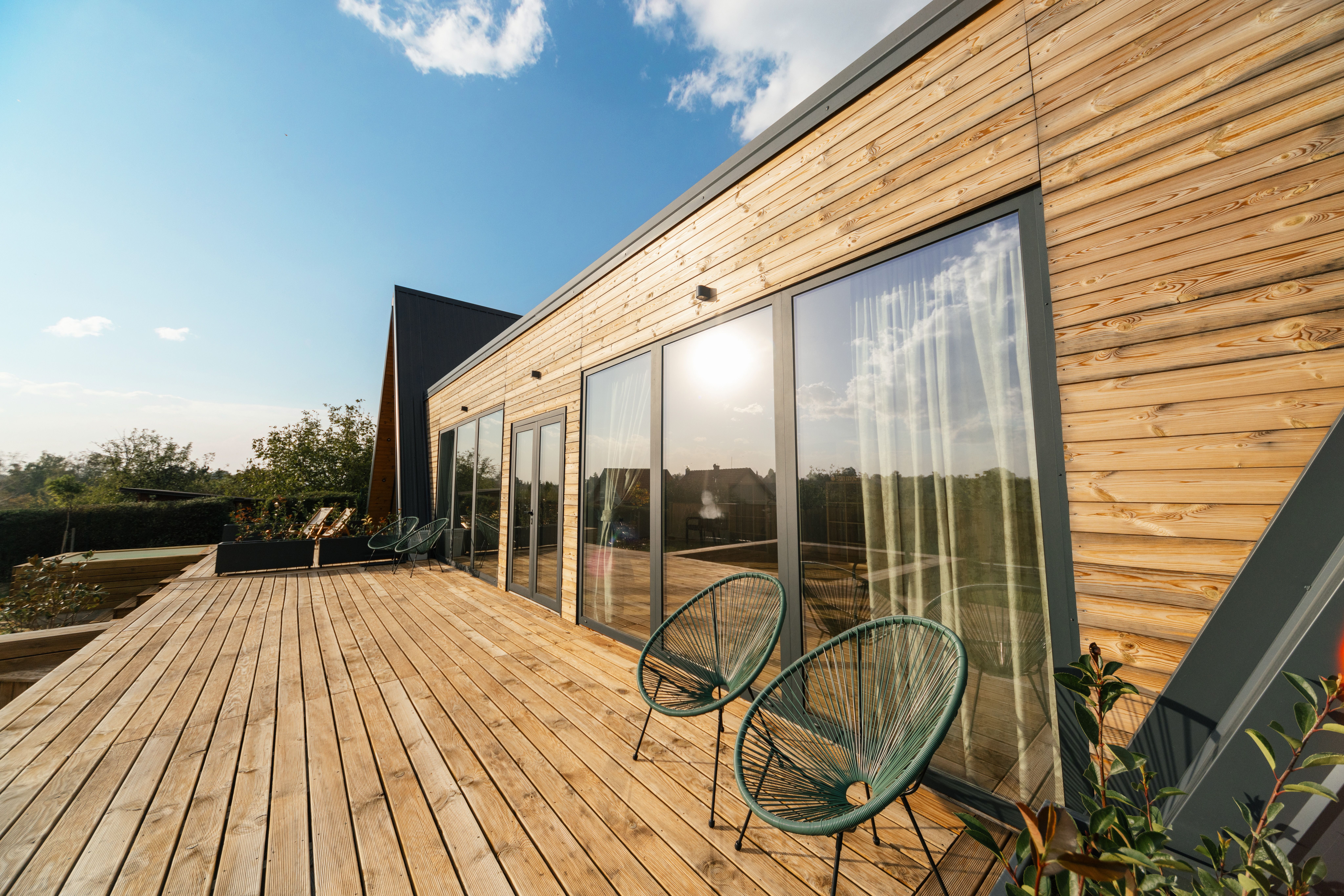 A modern house with wooden siding and large glass doors opening onto a spacious wooden deck with two patio chairs.