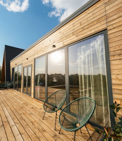 A modern house with wooden siding, large glass doors, and a wooden deck with two chairs.