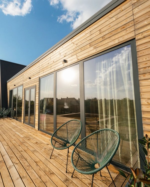 A modern house with wooden siding and large glass doors opening onto a spacious wooden deck with two patio chairs.