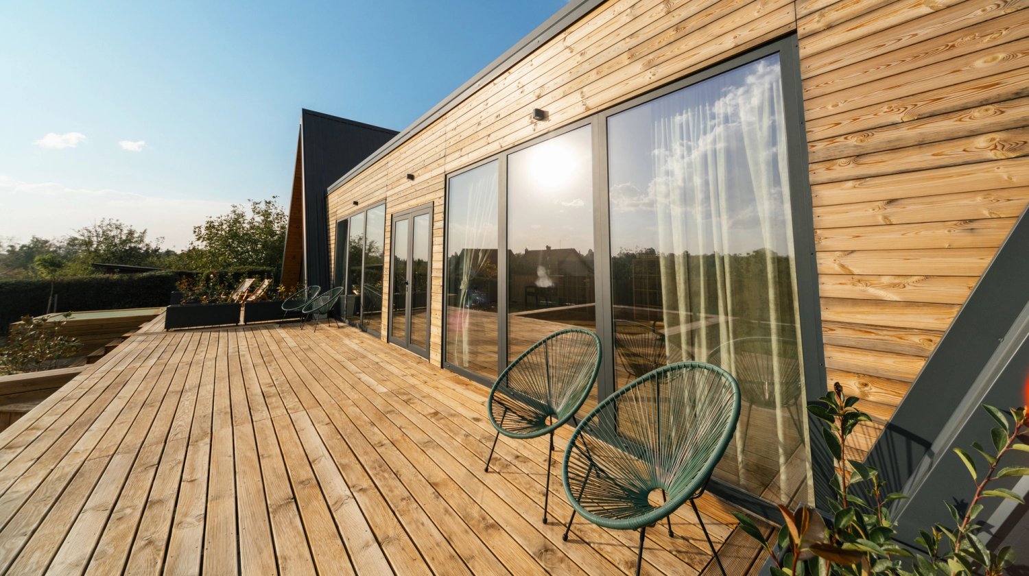 A modern house with wooden siding and large glass doors opening onto a spacious wooden deck with two patio chairs.