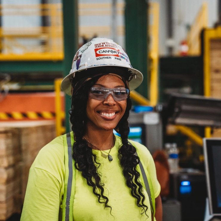 a woman wearing a hard hat and safety glasses is smiling in a factory .