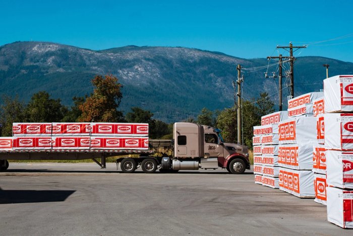 a semi truck is carrying a trailer with stacks of Canfor lumber out of a lumber yard.