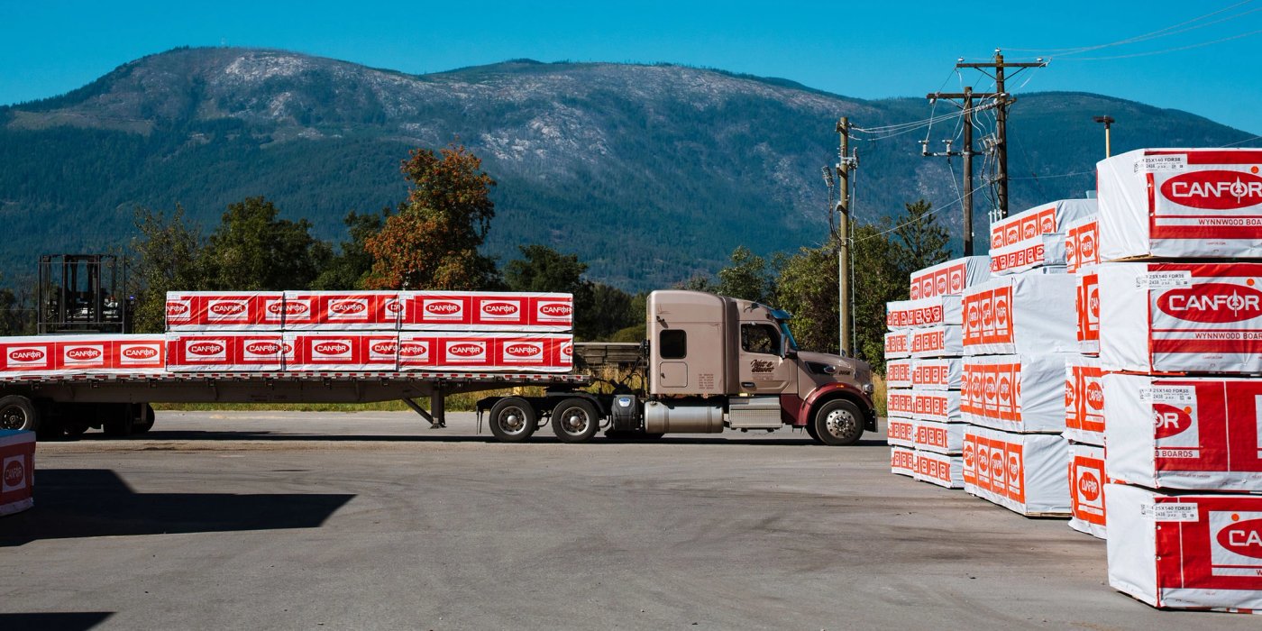 a semi truck is carrying a trailer with stacks of Canfor lumber out of a lumber yard.