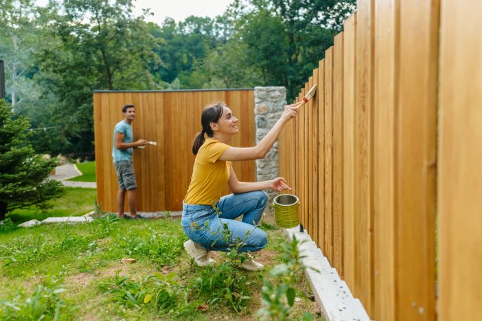 A smiling woman and man applying a finish to a wooden fence outdoors.