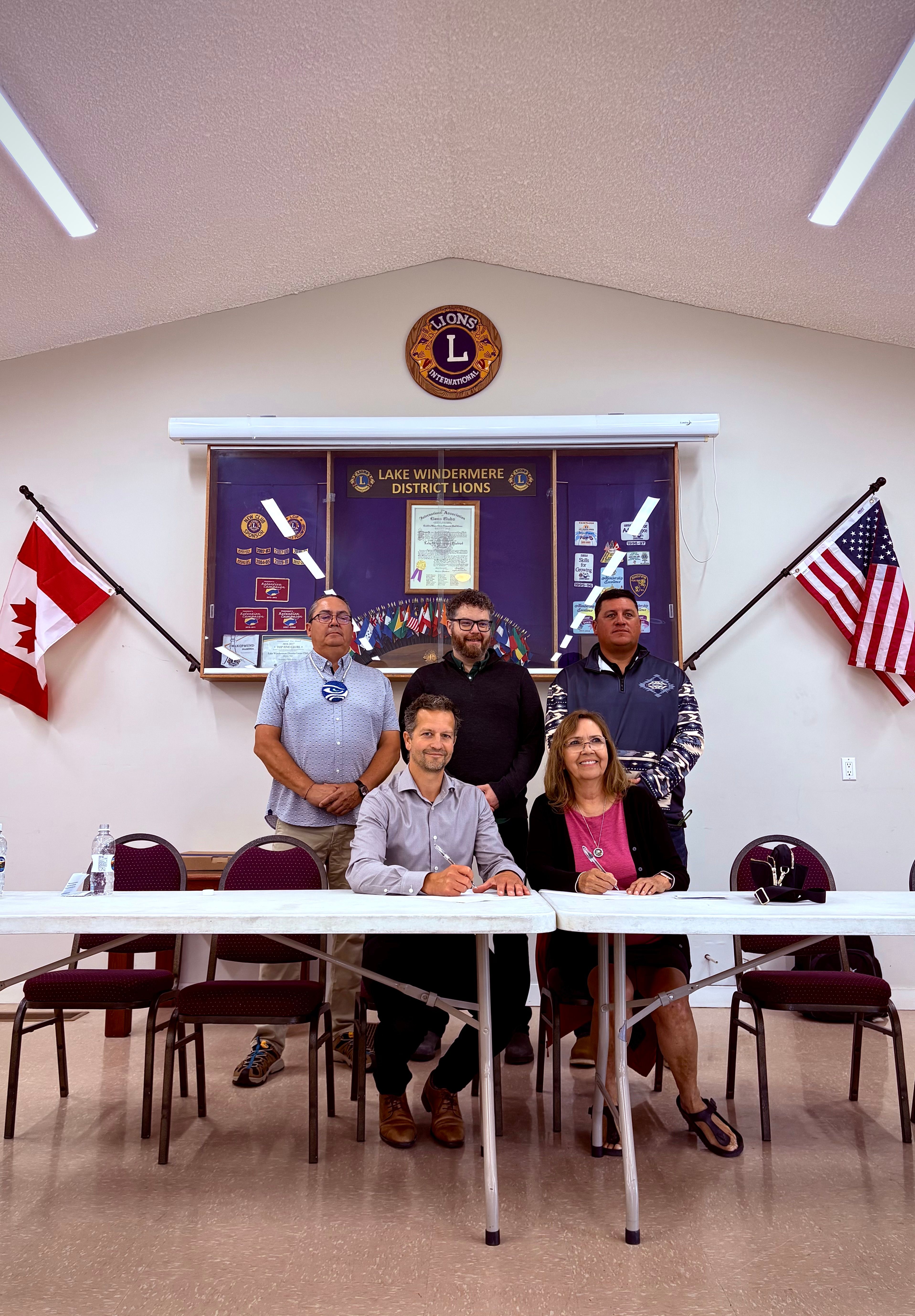 Two people sign documents at a table, while three others stand behind them, with banners and flags in the background.