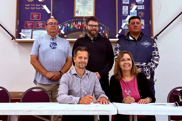 Two people sign documents at a table, while three others stand behind them, with banners and flags in the background.