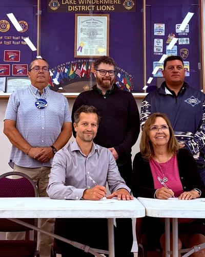 Two people sign documents at a table, while three others stand behind them, with banners and flags in the background.