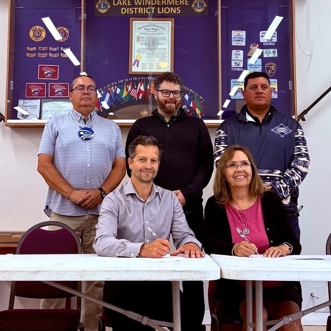 Two people sign documents at a table, while three others stand behind them, with banners and flags in the background.