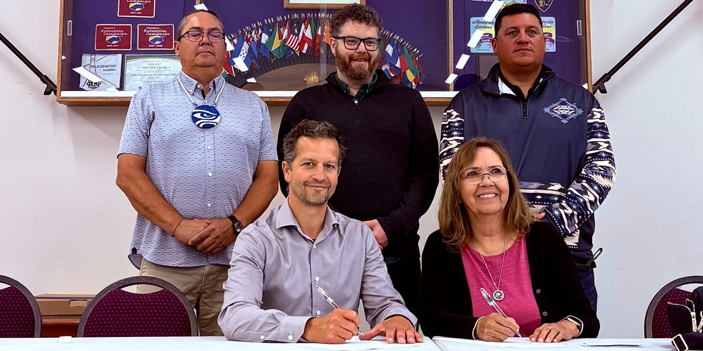 Two people sign documents at a table, while three others stand behind them, with banners and flags in the background.