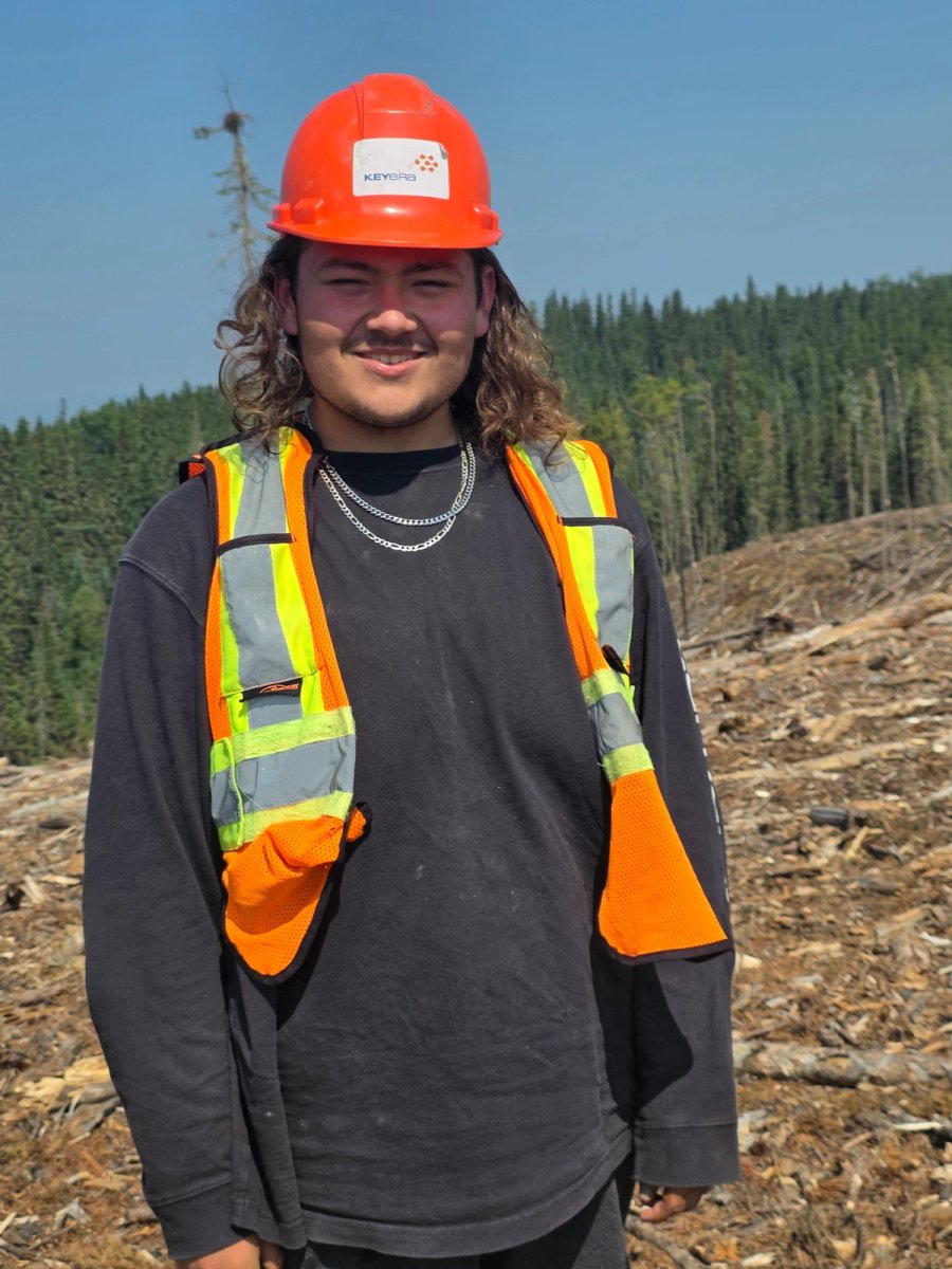 A smiling young man in an orange hard hat with a KEYOFED logo and a reflective safety vest stands in a logged forest clearing.
