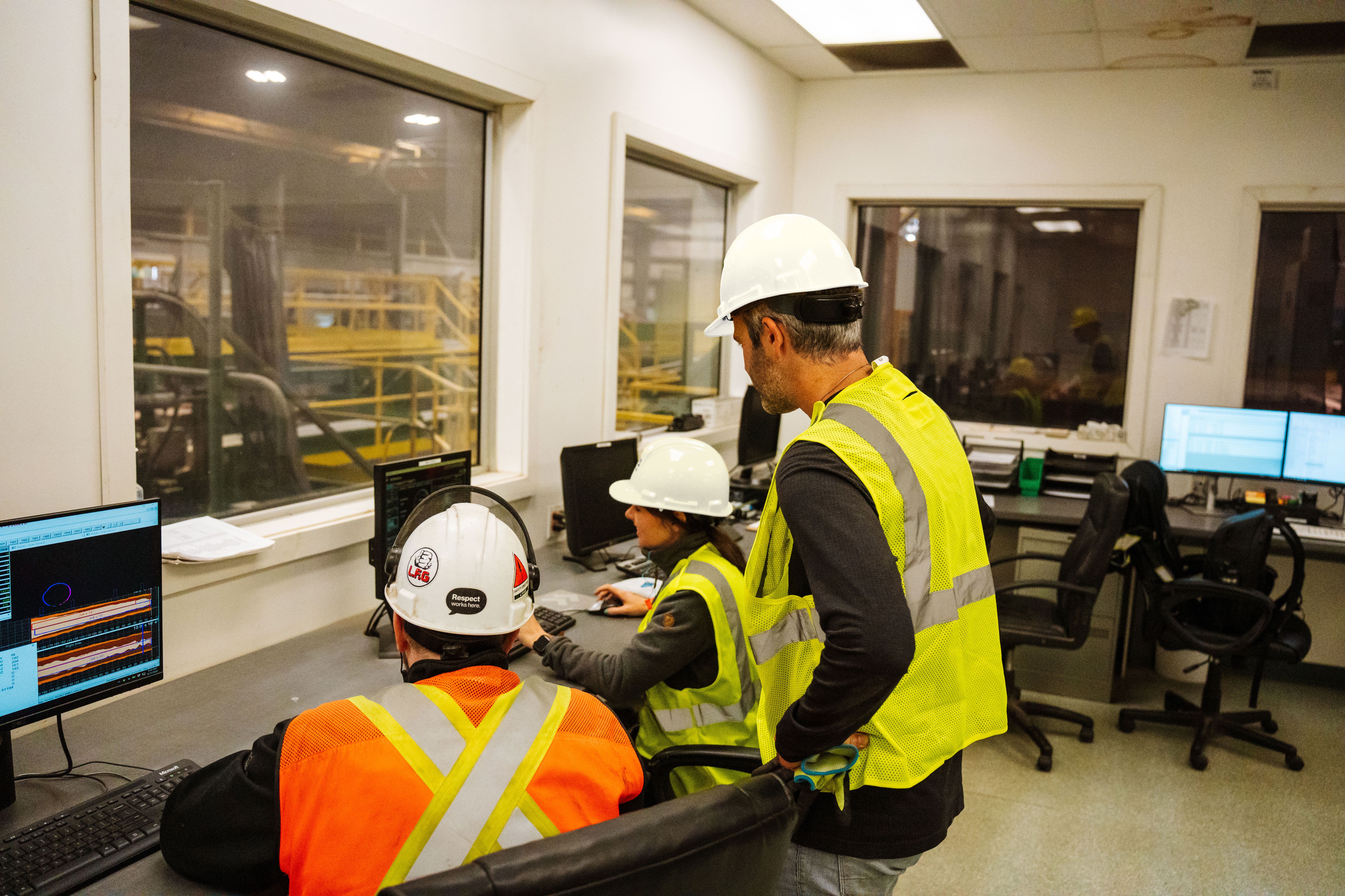 Three workers in hard hats and safety vests operate computers in an industrial control room with windows looking into a factory.