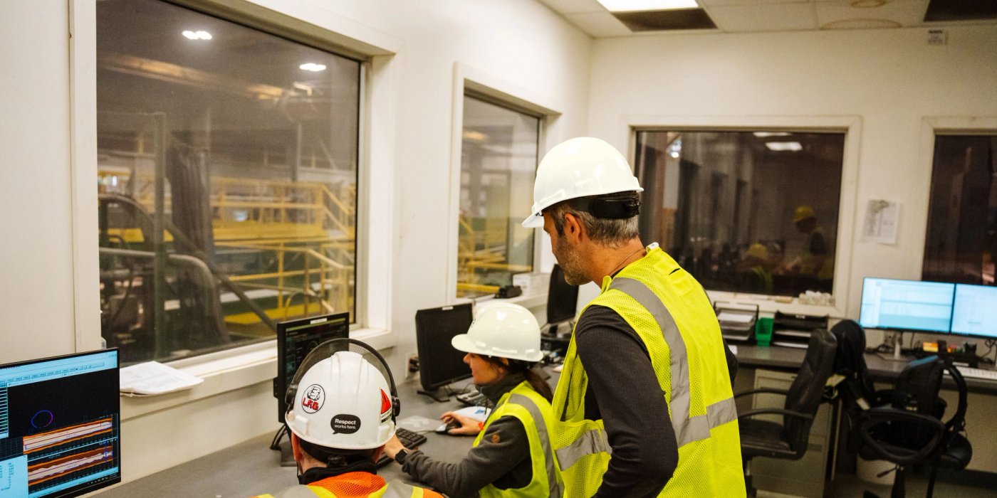 Three workers in hard hats and safety vests operate computers in an industrial control room with windows looking into a factory.