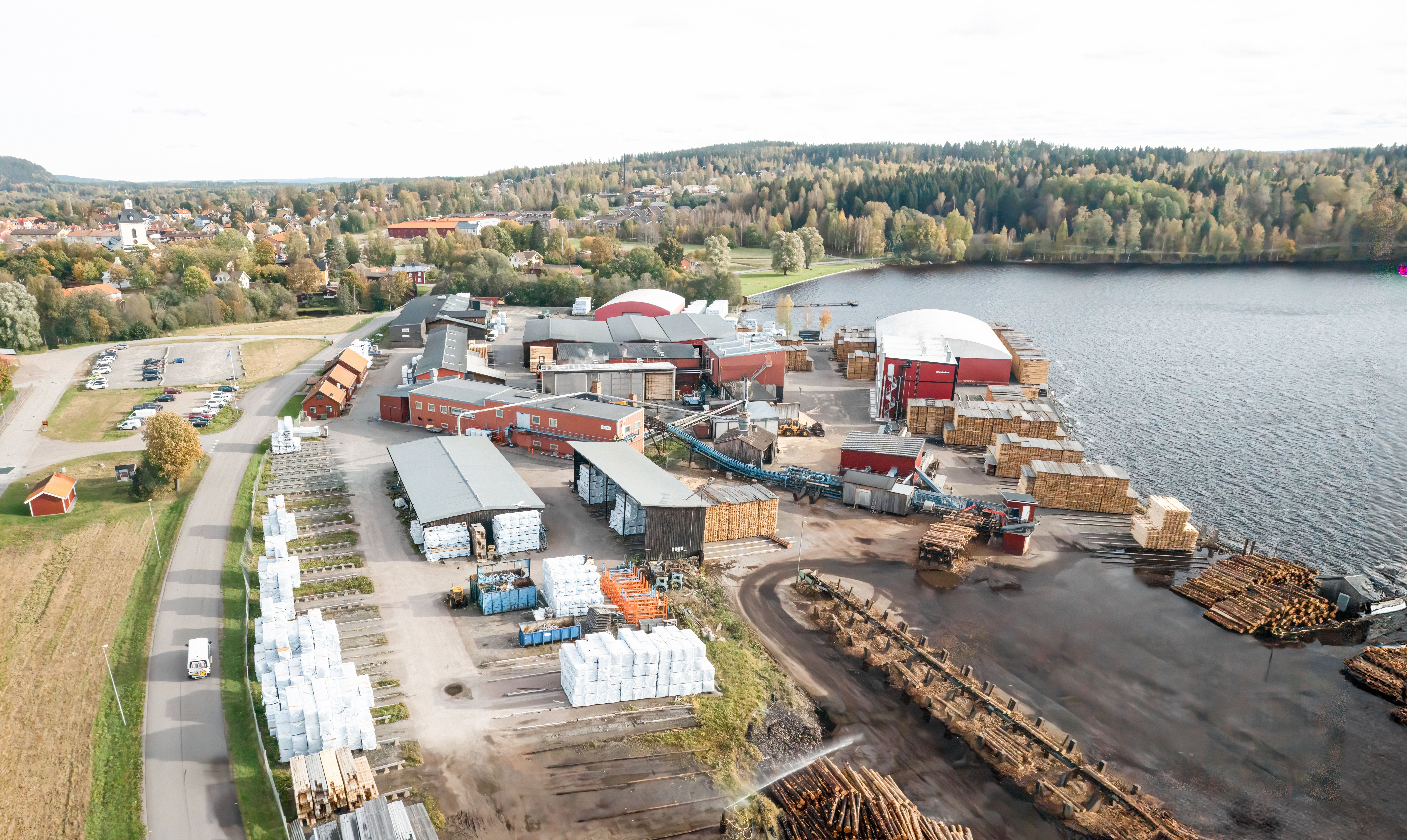 Aerial view of a timber processing plant on a lake, with stacks of logs and lumber, industrial buildings, and a town with forests in the background.
